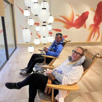 Two people sit in lounge chairs in hotel lobby. White lanterns hang from ceiling and large gold fish are painted on back wall.