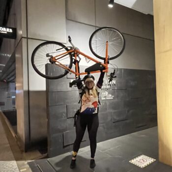 Cyclist holds orange bike above her head in front of hotel.