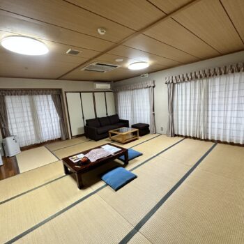 Large Japanese room with tatami floors. A couch and table are in one corner and a table with blue pillows for sitting is in the middle of the room.