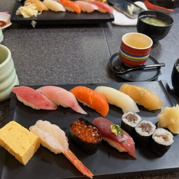 Rectangular plate filled with sushi and white pickled ginger. There are two tea cups and two bowls of miso soup also shown.