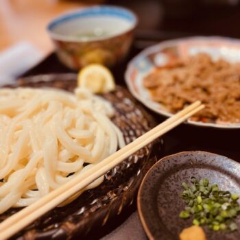 Cold Udon noodles are on brown basket with chopsticks sitting on the edge of the plate. Green onion on a smaller plate is next to the noodles and two other dishes are blurred in the background.