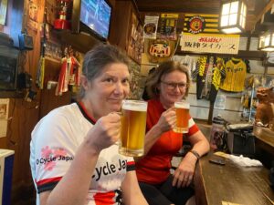 Two cyclists lift large glasses of draft beer in a busy bar in Japan. Wearing O cycle Japan jerseys.