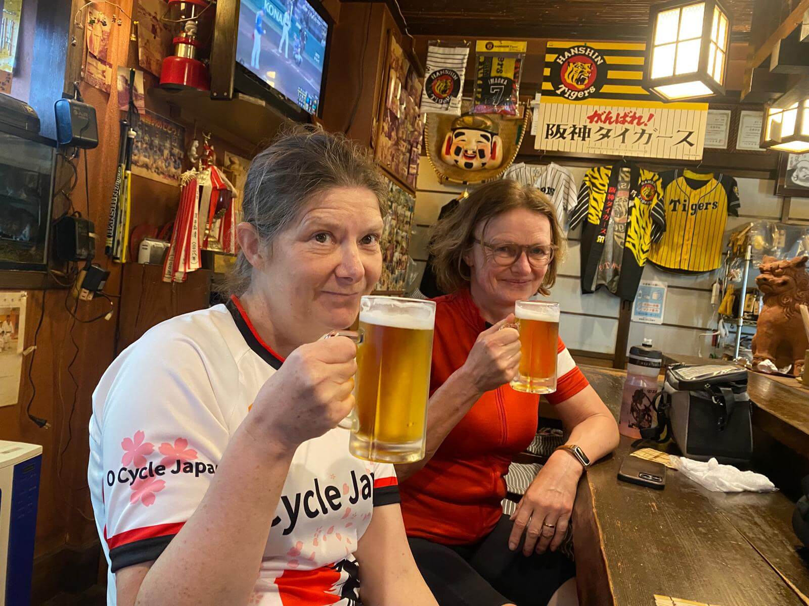 Two cyclists lift large glasses of draft beer in a busy bar in Japan. Wearing O cycle Japan jerseys.