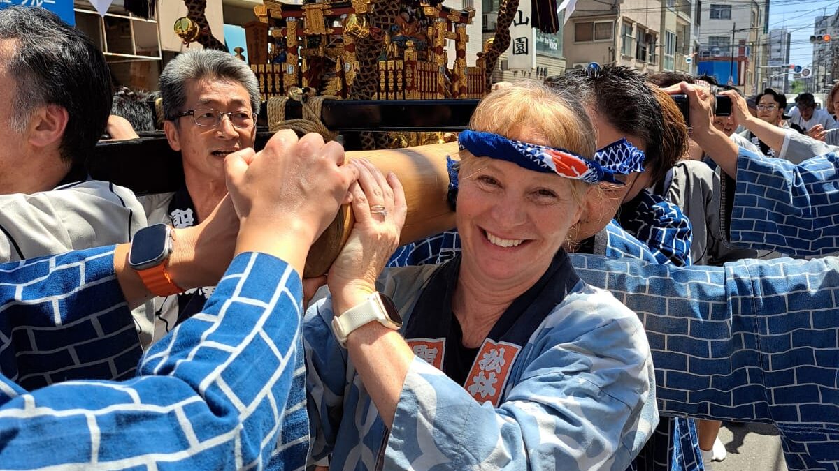 Group of people in blue jackets carry a Japanese float festival of gold and wood. Girl in front smiles broadly.