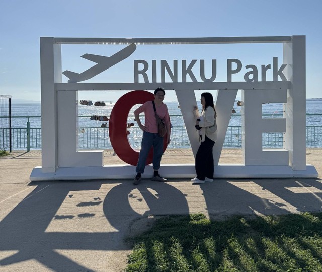 Couple stands in front of Rinku Park Love sign on a blue sky day with ocean in the background.