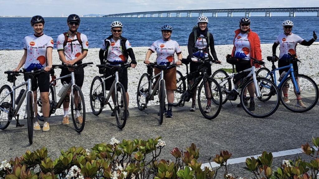 Group of seven cyclists pose on the promenade with Marble Beach, the ocean and the long bridge to the Kansai International Airport in background.
